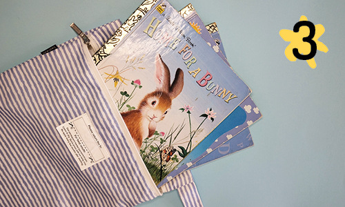 A blue and white striped wet bag, repurposed as a library bag. The bag is unzipped to reveal 3 Golden Books inside, the front book is titled Home For a Bunny with an illustrated rabbit on the cover.