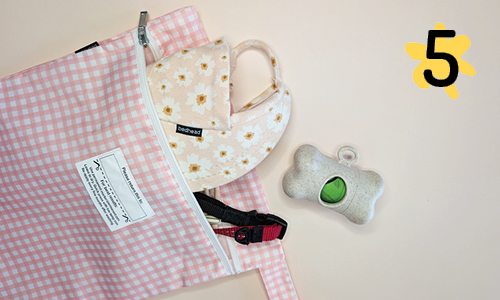 A pink wet bag with a gingham patterned design, repurposed as a pet accessory holder. The bag is unzipped to reveal a Bedhead Hats dog hat, a sanitary bag holder in the shape of a bone, and a red collar.