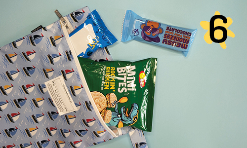 A blue wet bag with a sailing boat patterned design, repurposed as a snack bag. The bag is unzipped to reveal two chocolate bars in blue packaging and a green bag of rice wheel snacks. 