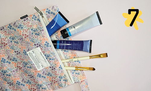 A pink, blue and purple floral wet bag, repurposed as a craft supply bag. The bag is unzipped to reveal 3 tubes of acrylic paint in blue colours, and two white paint brushes.